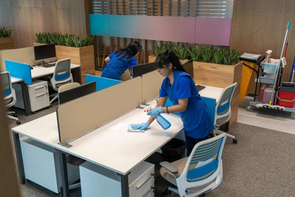 Two women in blue shirts and cleaning gloves cleaning desks in an El Paso office.