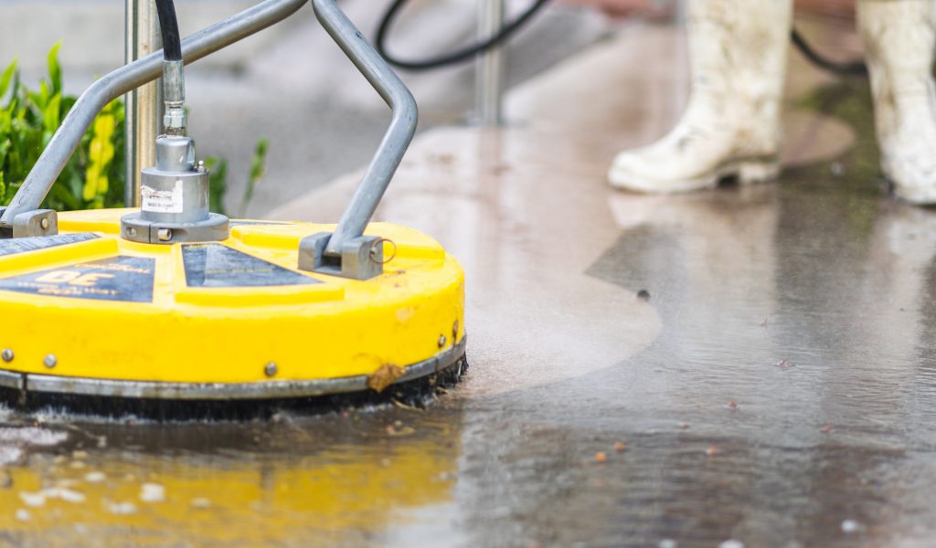 A worker using industrial equipment to perform pressure washing services on a sidewalk.